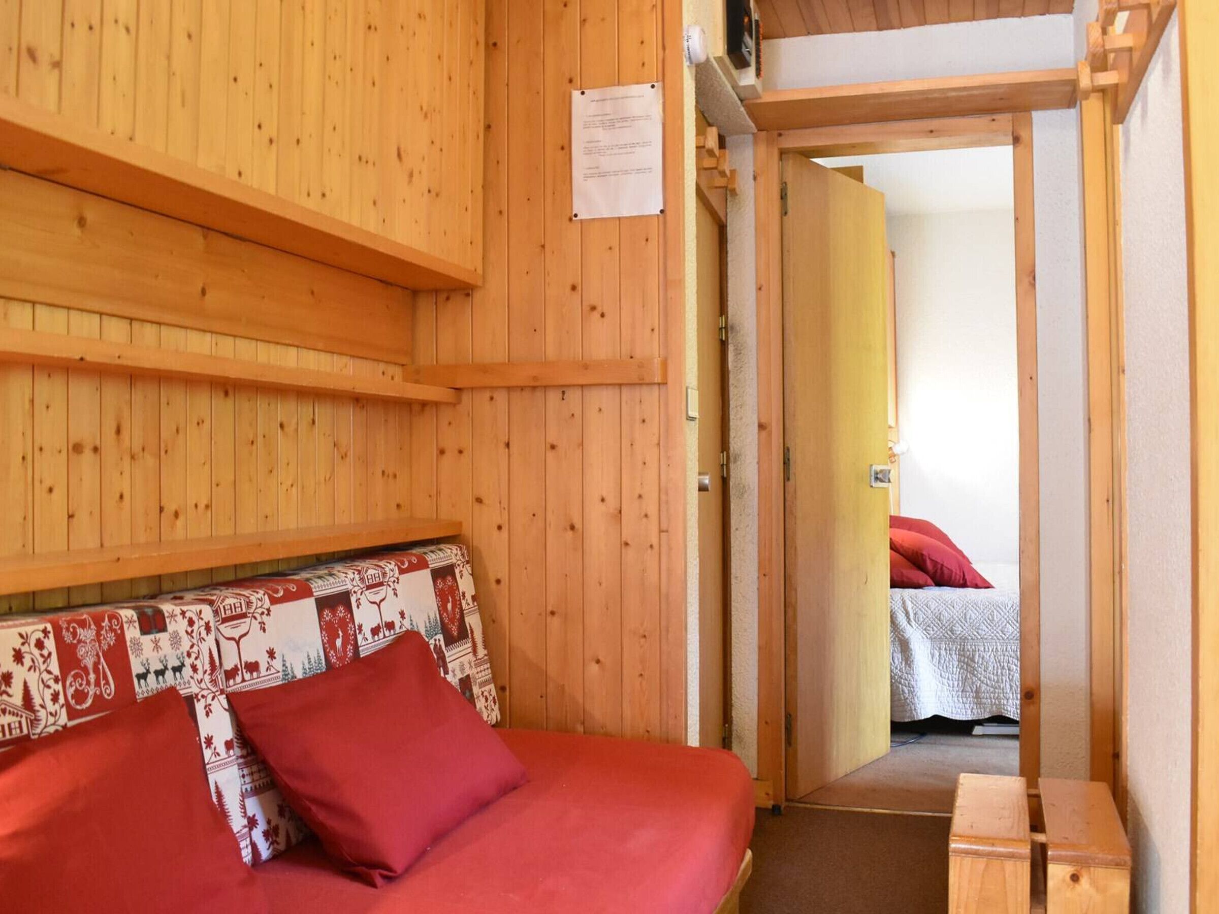 A wooden room with a bed, a bench, and a door leading to another room.