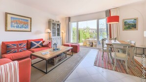 A modern living room with a sofa set, a wooden coffee table, a dining table with chairs, a cabinet, and a large window offering a view of the outdoors.