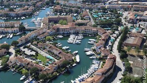 An aerial view of a marina with numerous boats docked, surrounded by buildings with terracotta roofs.