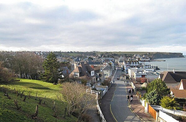 A coastal town with houses along a curved road, a clear view of the sea, and a hilly landscape in the background.
