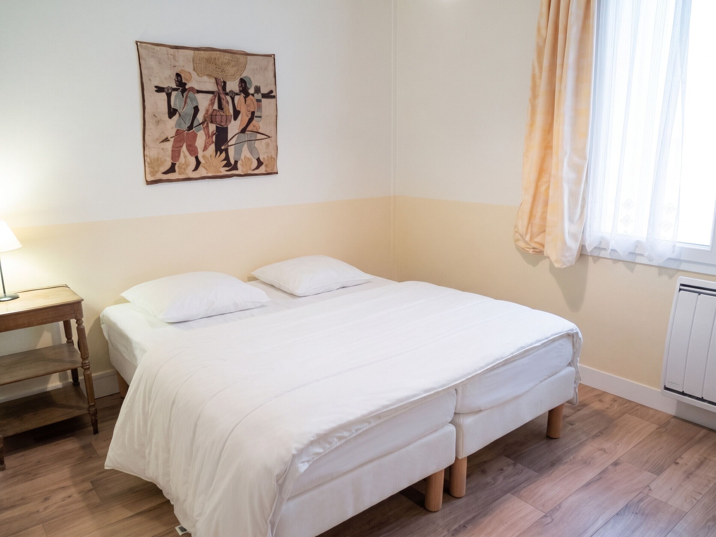 Een slaapkamer met een bed, nachtkastje, lamp, schilderij en een raam met gordijnen.