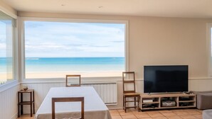 A room with a large window overlooking a beach, a bed, a TV, and a seating area. - Homerez - House in Arromanches-les-Bains (Arromanches-les-Bains)
