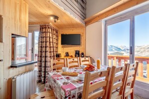 Une pièce chaleureuse avec un décor intérieur en bois, un téléviseur fixé au mur, une table à manger avec des chaises, et une vue sur les montagnes par de grandes fenêtres.