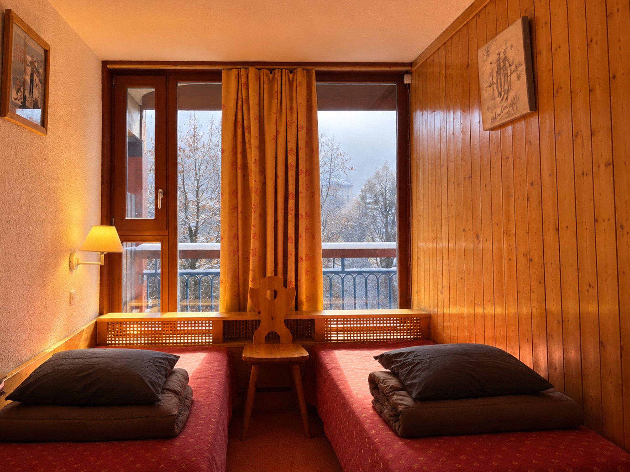 Une pièce avec deux lits, un banc en bois, une fenêtre avec des rideaux, une lampe et un tableau encadré au mur.