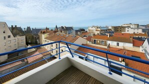 A rooftop terrace with a wooden floor and blue railings, overlooking a coastal cityscape with terracotta-roofed buildings and a clear sky.