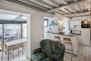 Une cuisine moderne avec des meubles blancs, un four à micro-ondes et un petit îlot central. On y trouve un coin repas avec une table et des chaises, ainsi qu’une grande fenêtre donnant sur un mur en pierre.