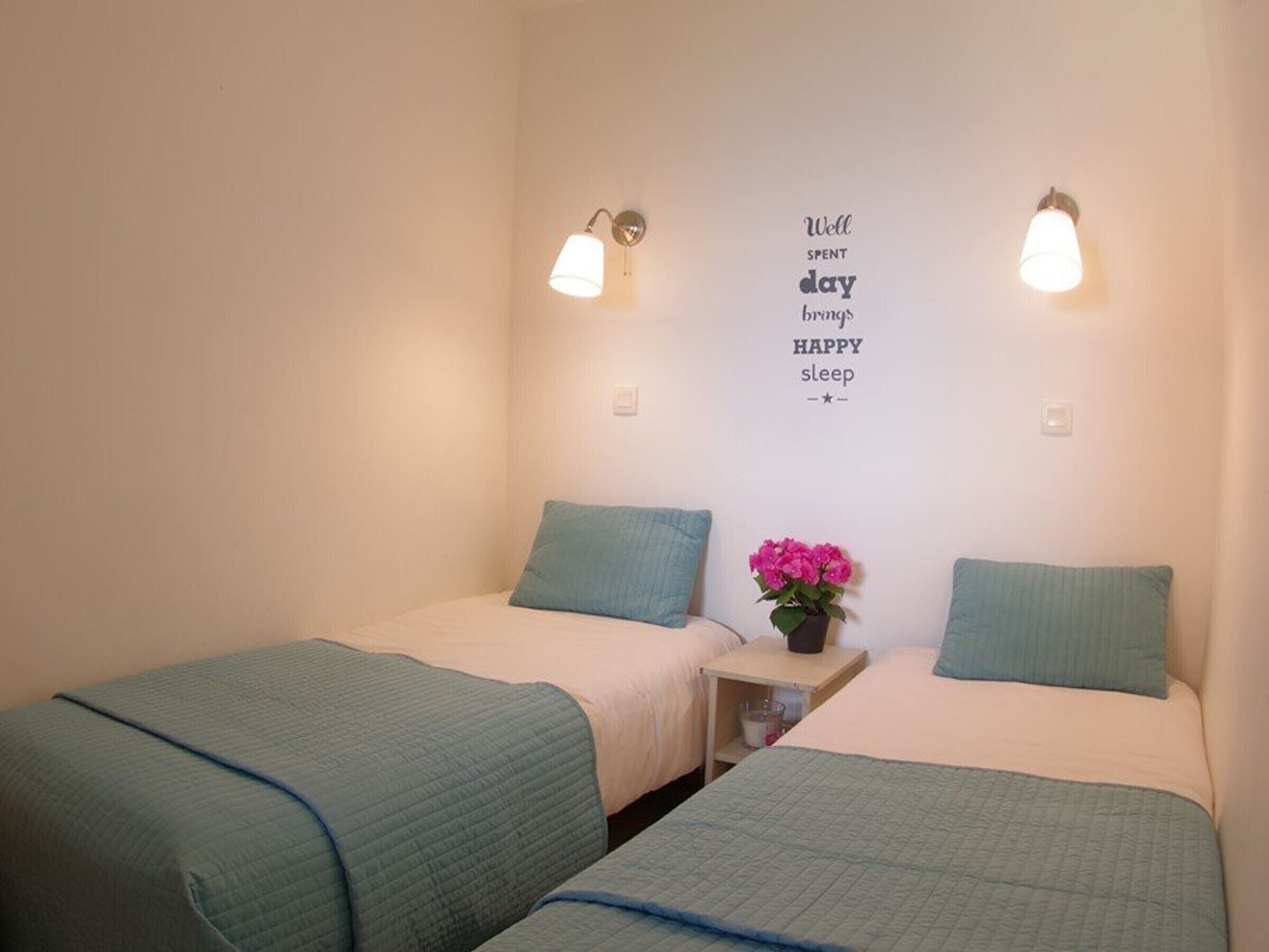 A twin room with two beds, each with a blue quilted bedspread and matching pillows. There is a wall-mounted light fixture on each side of the bed, and a wall decoration with a motivational quote.