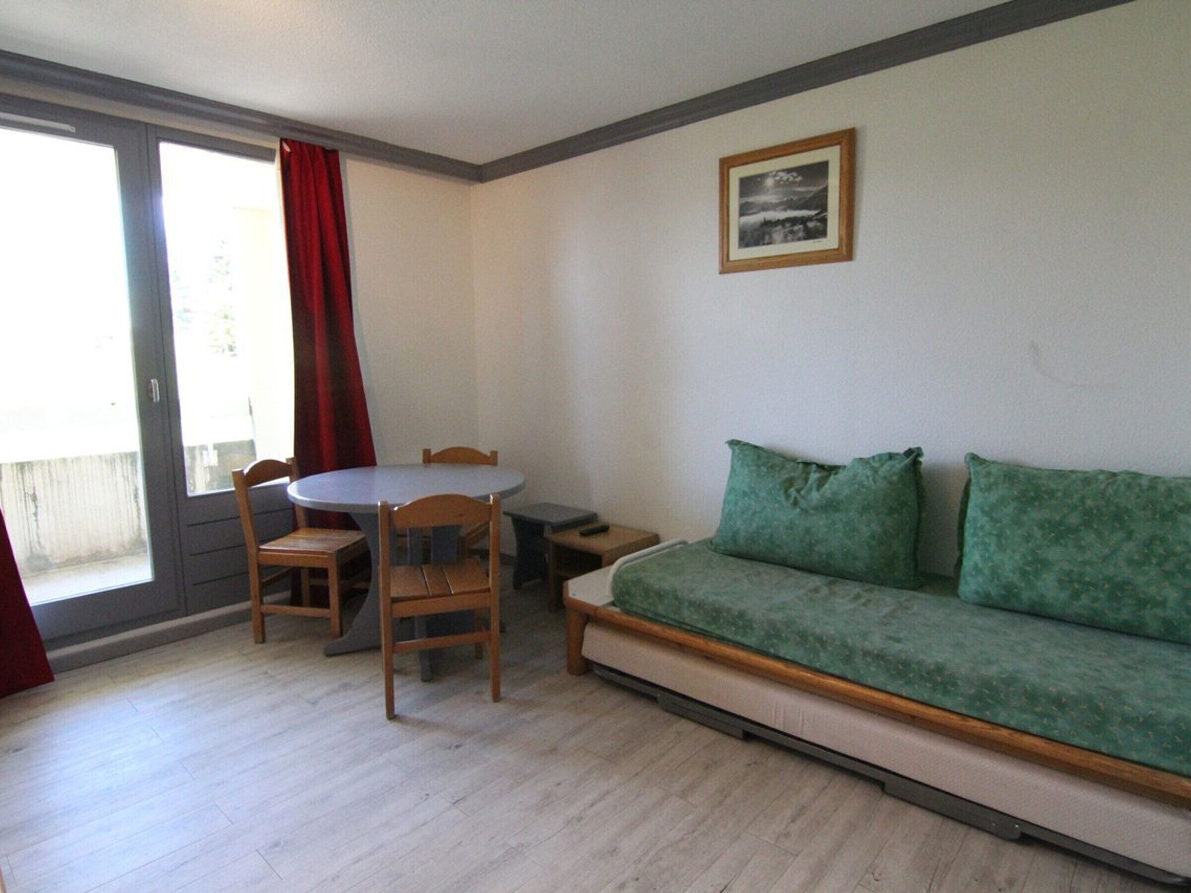 Ein Zimmer mit einer Couch, einem Esstisch mit Stühlen, einem kleinen Beistelltisch und einer Glasschiebetür mit rotem Vorhang.