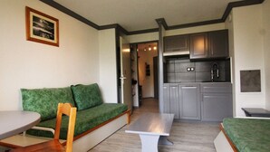 A compact living space with a green sofa, a wooden chair, a small dining table, and a kitchenette with grey cabinets and a stainless steel sink.