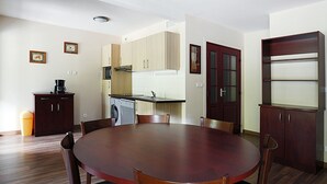 A dining area with a round wooden table and chairs, a kitchen with cabinets and appliances, and a trash can.