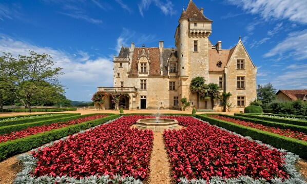 Ein großes, historisches Gebäude mit einer symmetrischen Fassade, einem zentralen Innenhof mit einem Brunnen und einem formell gestalteten Garten mit roten Blumen.
