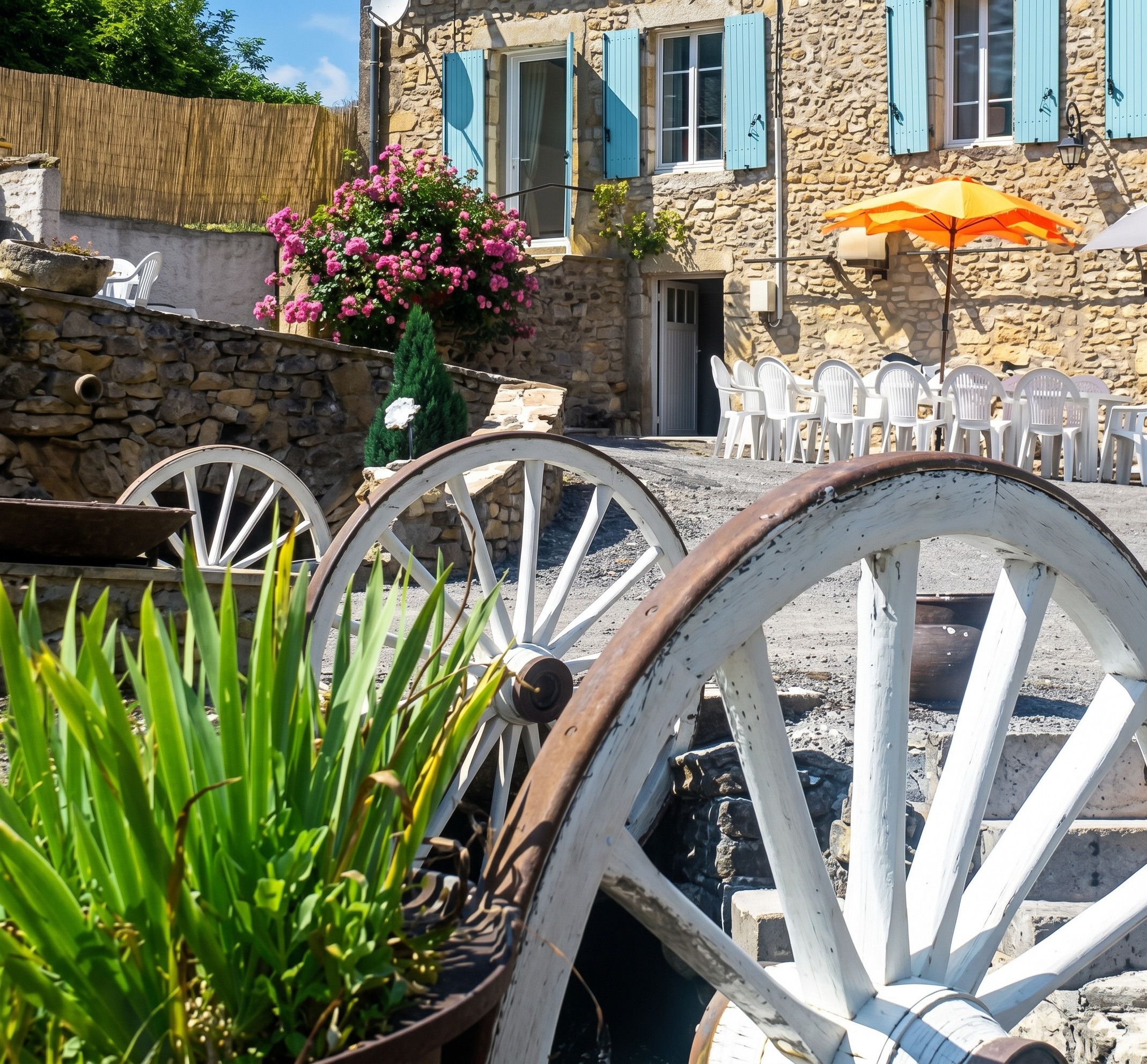 Sitzgelegenheit im Freien mit weißen Stühlen und einem Tisch, ein Steinhaus mit blauen Fensterläden und ein Blumentopf mit rosa Blumen.