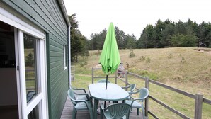 Eine Terrasse mit Tisch und Stühlen, einem grünen Sonnenschirm und Blick auf ein Wiesenfeld mit Bäumen.