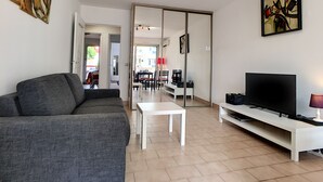 Una sala moderna con un sofá gris, una mesa de centro blanca, un televisor de pantalla plana sobre un soporte blanco y una puerta corrediza de vidrio que da a un comedor.