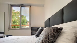 Una cama bien hecha con almohadillas estampadas, una ventana con vistas a árboles y una mesita de noche con una lámpara.
