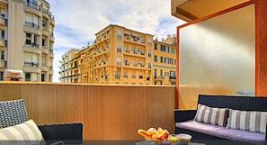 A balcony with wicker furniture, a glass table, and a view of a yellow building.