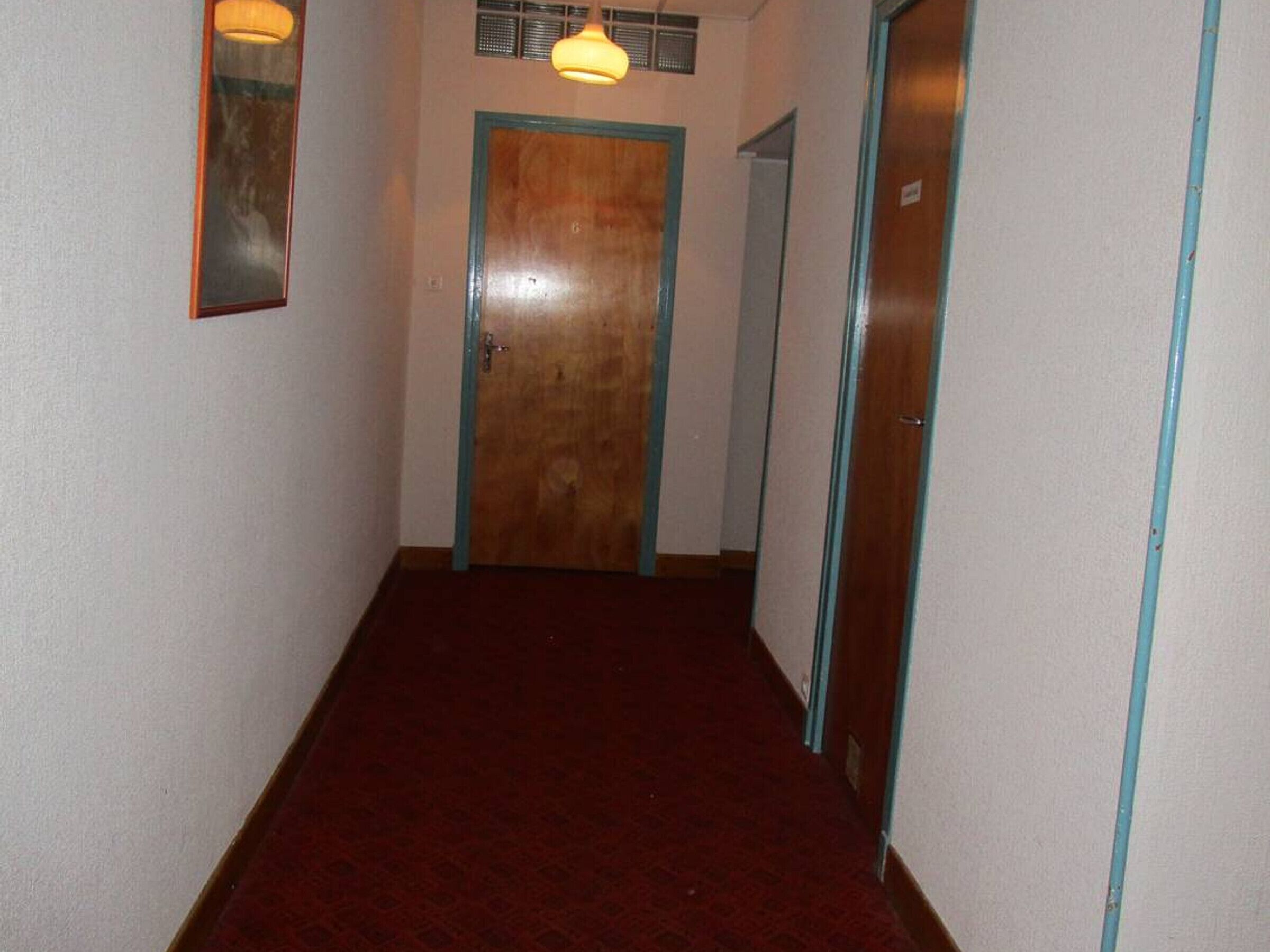 A narrow hallway with a wooden door at the end, a mirror on the left, and a light fixture above.