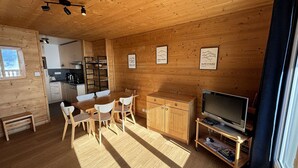 Interior de uma cabana de madeira com sala de jantar, cozinha e uma TV sobre um carrinho.