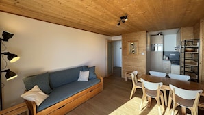 Uma sala de estar acolhedora com um sofá, uma mesa de jantar de madeira e cadeiras. Uma cozinha com eletrodomésticos modernos pode ser vista ao fundo.