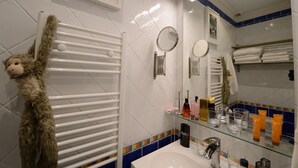 A bathroom with a white tiled wall, a glass shelf with toiletries, a white sink, and a monkey decoration. - A2DF Boogie-Woogie (París)