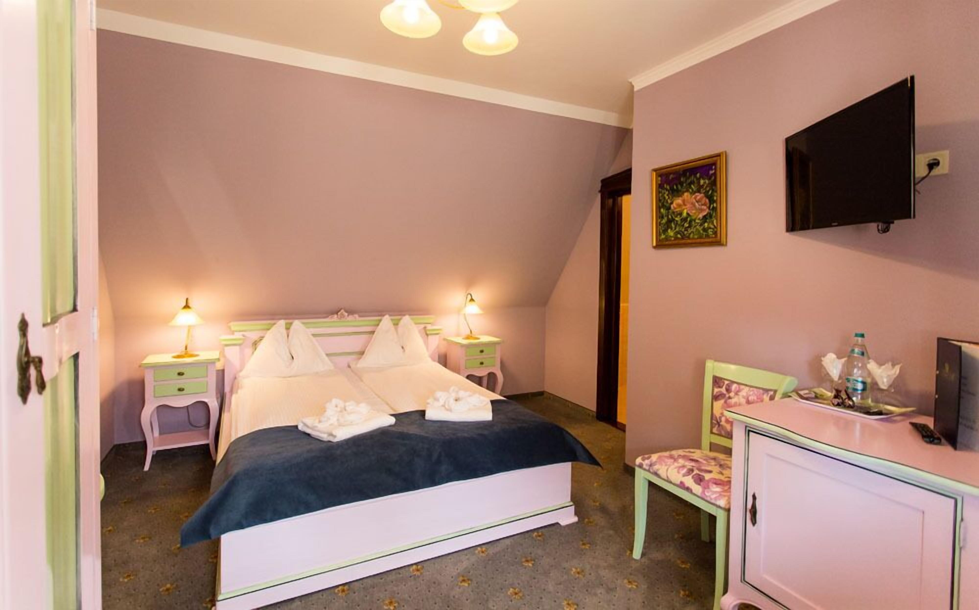 Ein Schlafzimmer mit Bett, Nachttischen, einem Schminktisch und einem Fernseher. An der Wand hängt ein Gemälde.