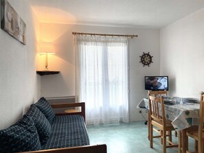 A room with a bed, a TV, a table with chairs, and a window with curtains.