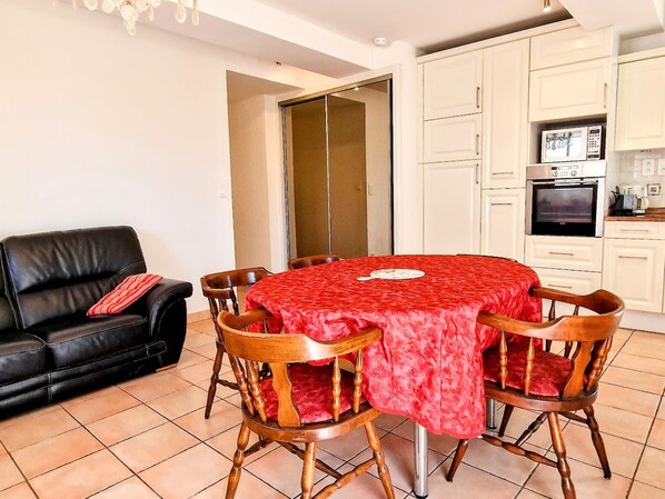 A dining area with a red tablecloth, wooden chairs, a black leather sofa, and a kitchen with white cabinets and appliances.