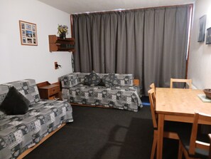 Een woonkamer met een bank, een eettafel en een stoel. Er staat een klein houten bijzettafeltje en een wandplank met een vaas.