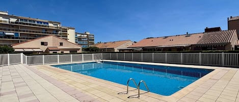 Ein rechteckiger Pool mit einer gefliesten Liegefläche, umgeben von einem weißen Zaun, und Gebäuden im Hintergrund.