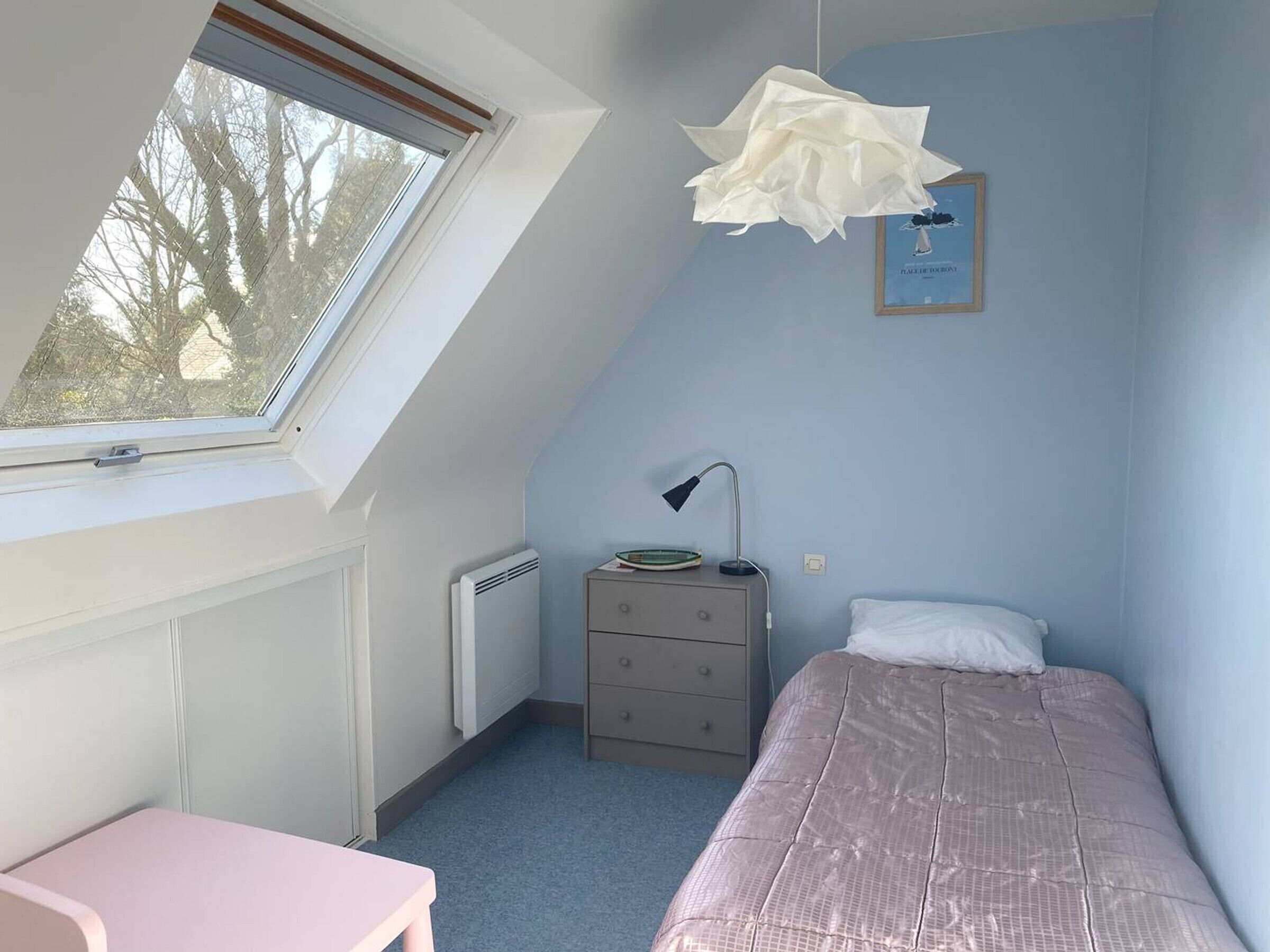Un dormitorio con techo inclinado, una ventana, una mesita de noche, una lámpara, una cama y un cuadro enmarcado en la pared.