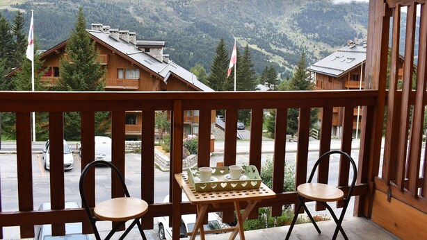A balcony with two chairs and a table, overlooking a mountainous landscape with buildings and flags. - Méribel: Cozy 2-Room Apt, 4 Pax, near Shops and Slopes (Méribel)