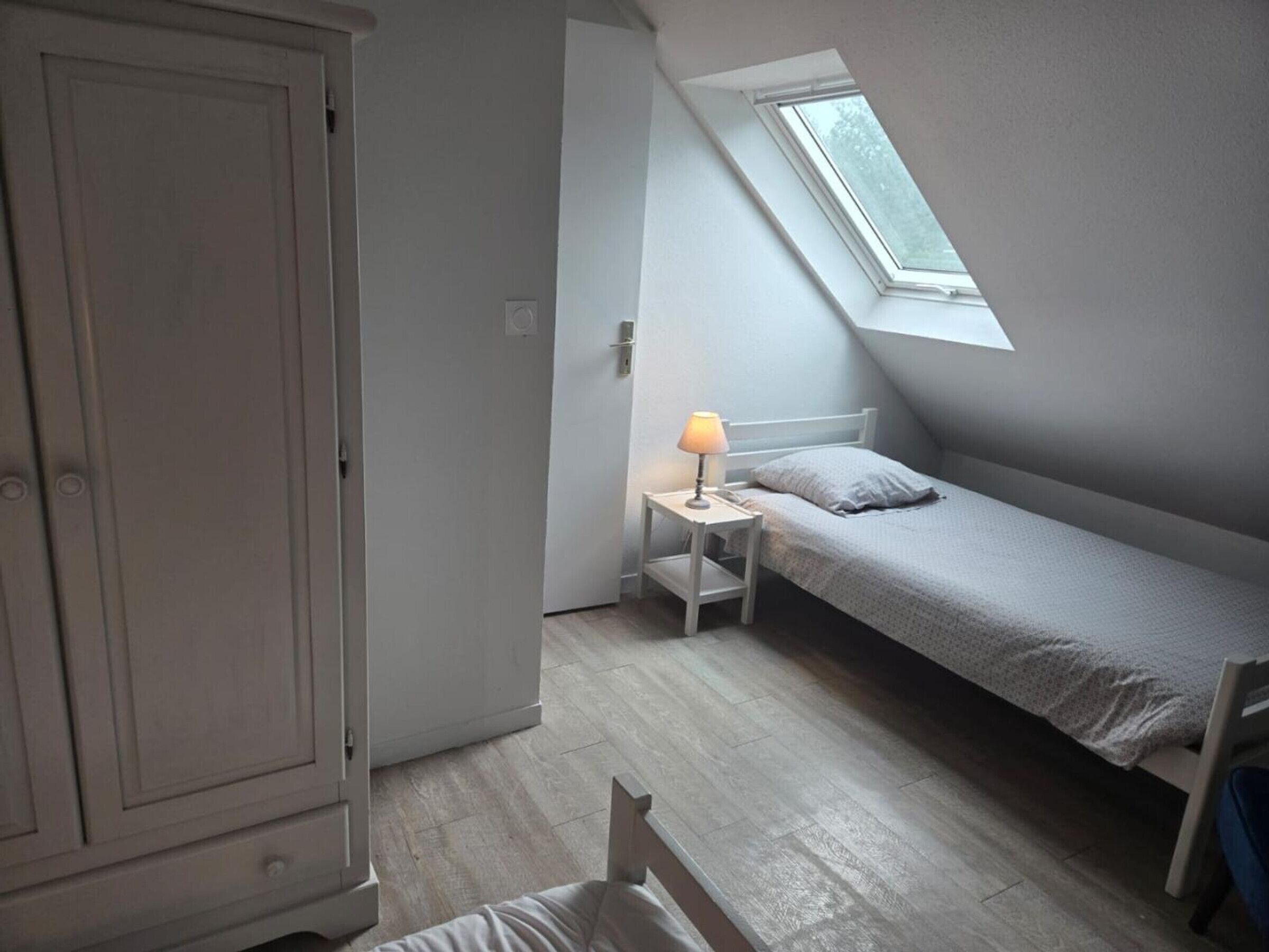 A small, single-bed room with a slanted ceiling and a skylight, a bedside table with a lamp, and a wardrobe.