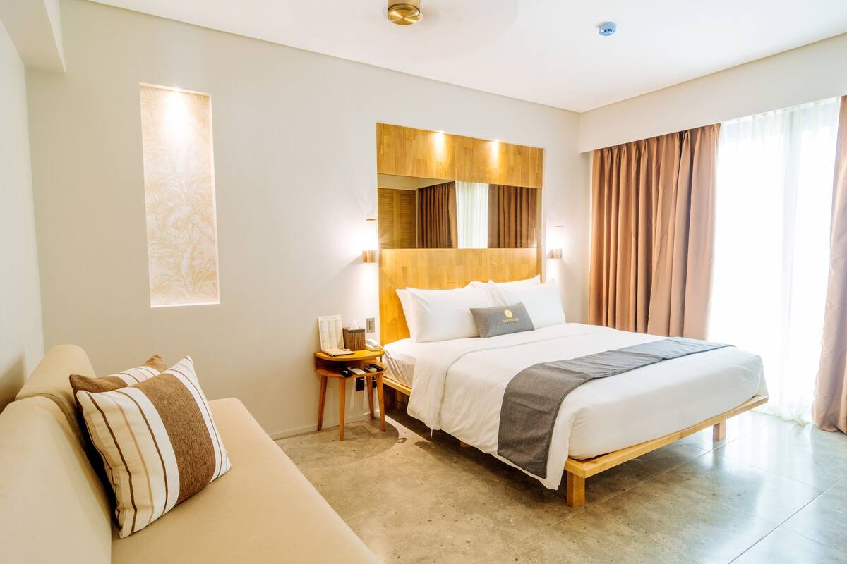 A hotel room with a bed, a nightstand, a lamp, a sofa, and a large window with curtains.