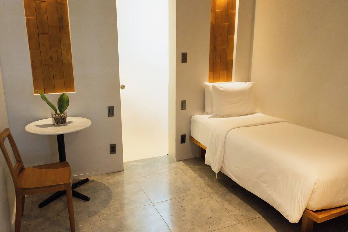 A small, clean room with a single bed, a chair, and a small table with a plant.