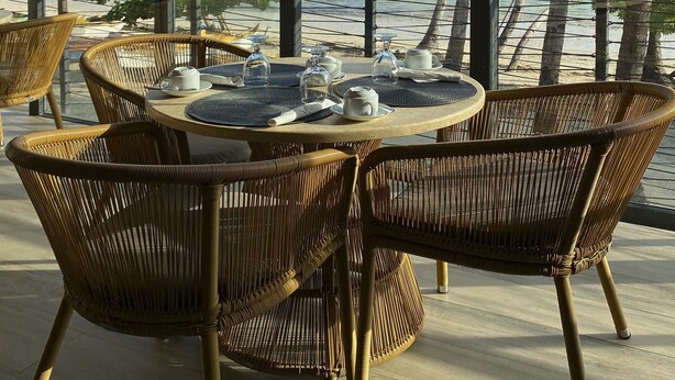 Een eethoek met rieten stoelen en een ronde tafel, met uitzicht op een strand met palmbomen en de zee.