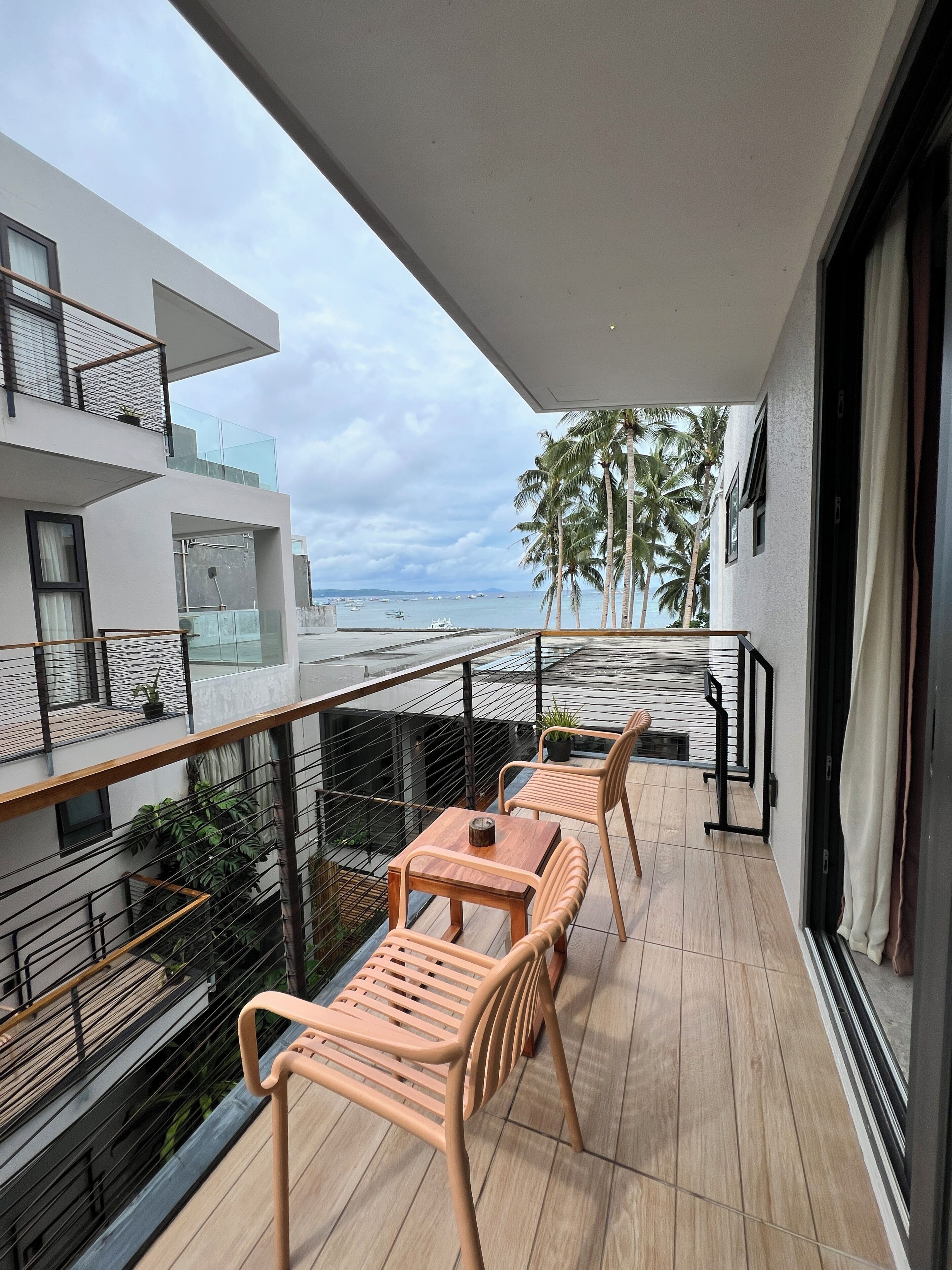 A balcony with two wooden chairs and a table, overlooking a beach with palm trees and a modern building.