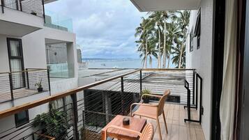 Un balcón con dos sillas de madera y una mesa, con vista a una playa con palmeras y un edificio moderno.