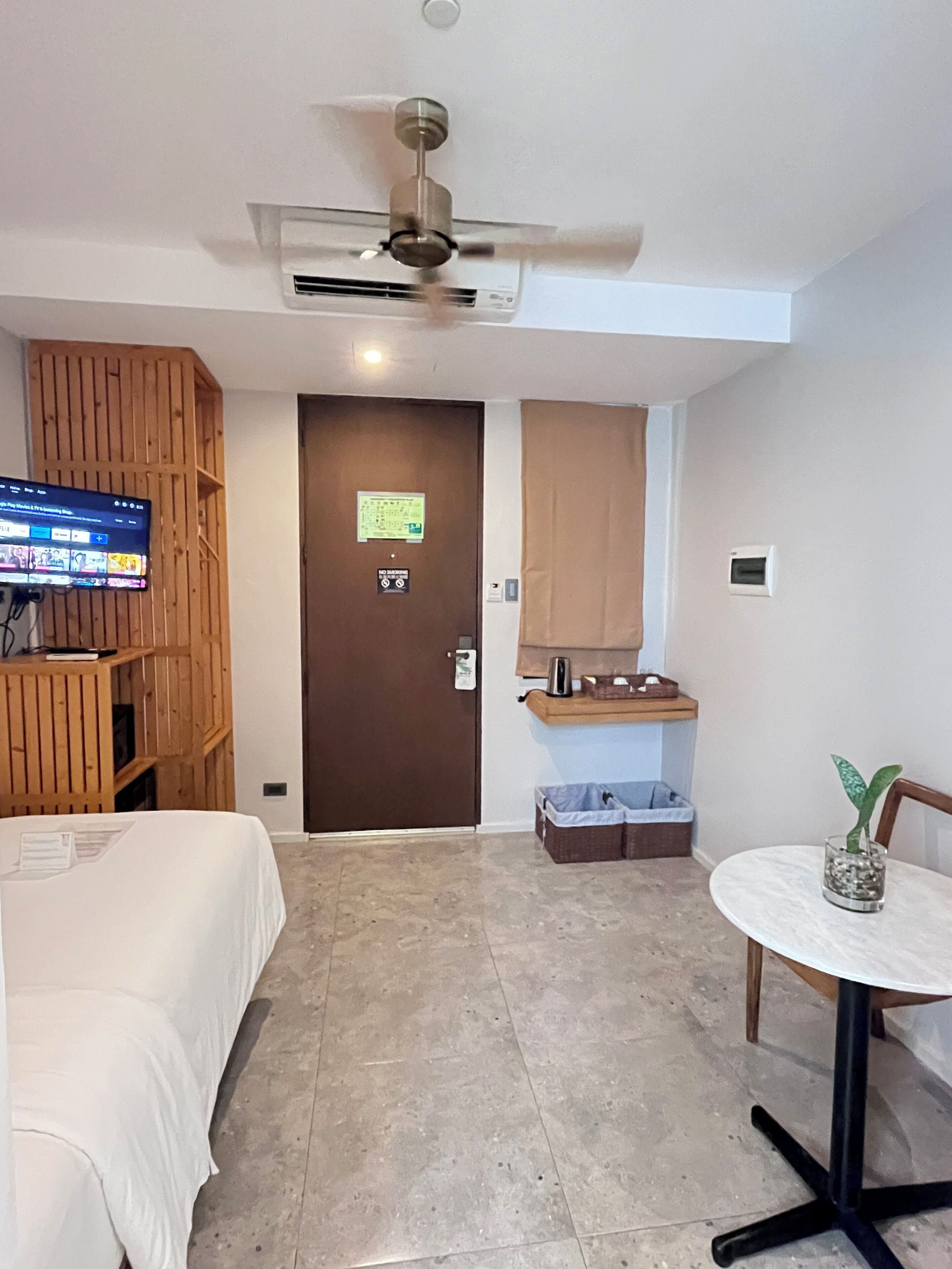 Ein Hotelzimmer mit einem Bett, einem Fernseher auf einem Holzboden, einem Deckenventilator, einer Tür mit einem Schild, einem kleinen Tisch mit einer Pflanze und einem Mülleimer.