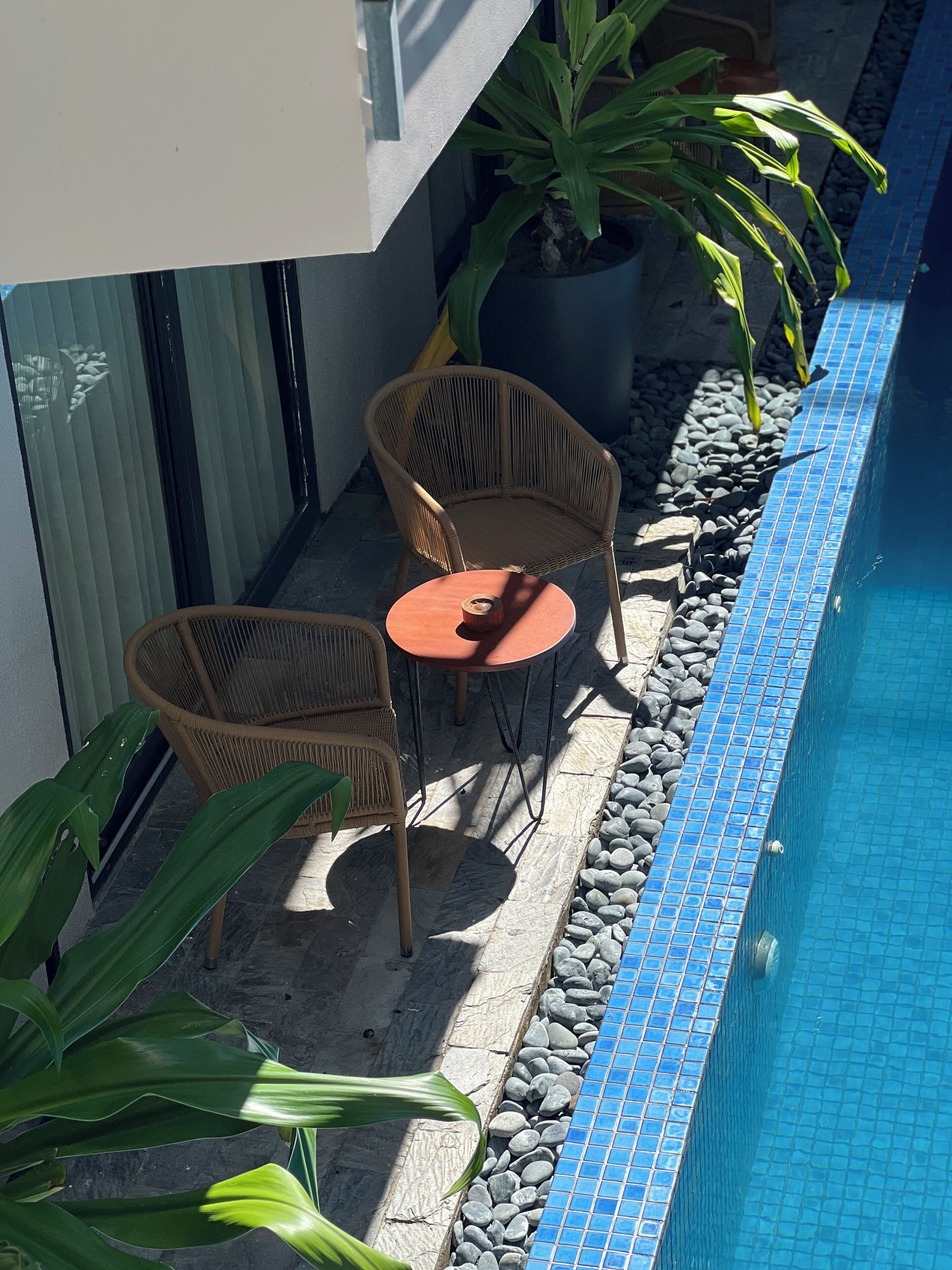 A poolside area with a small table and two wicker chairs, surrounded by potted plants and a tiled pool edge.