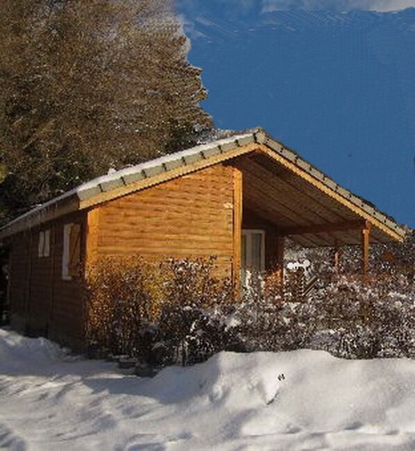 A wooden cabin with a sloped roof, surrounded by snow-covered ground and bushes.