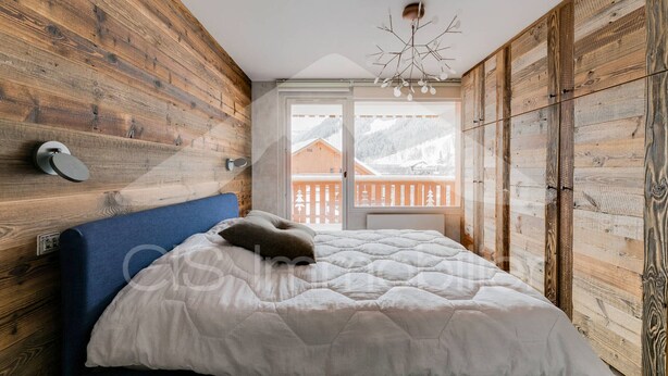 Un dormitorio con paredes de madera, una cama con colcha blanca, cabecera azul y vista a una montaña nevada a través de una ventana.