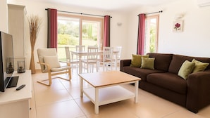 Ein Wohnzimmer mit braunem Sofa, weißen Sesseln, einem Holztisch und einem Fernseher auf einer weißen Kommode.