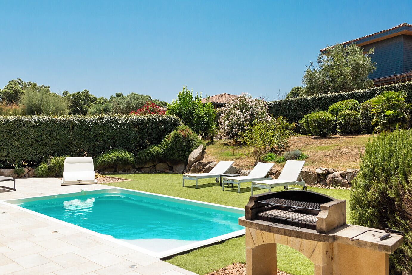 A pool with lounge chairs, a barbecue grill, and a well-maintained garden with various plants and a hedge.