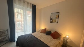 A hotel room with a bed, two bedside lamps, a window with curtains, and a framed picture on the wall. - Lovely 1BR in Arcachon - Parking, WiFi, Near Beach (Arcachon)