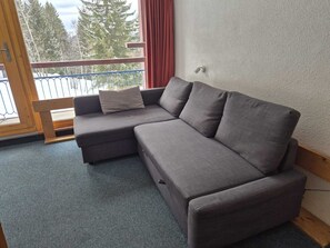 Ein graues Ecksofa mit einer Holzbank an einer Wand, ein Fenster mit Blick auf Bäume und ein Balkon mit Metallgeländer.