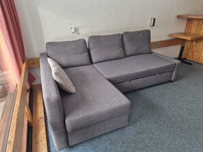 Ein graues Ecksofa mit einer Holzbank links, einer weißen Wand mit Struktur und einem kleinen Holzregal rechts.