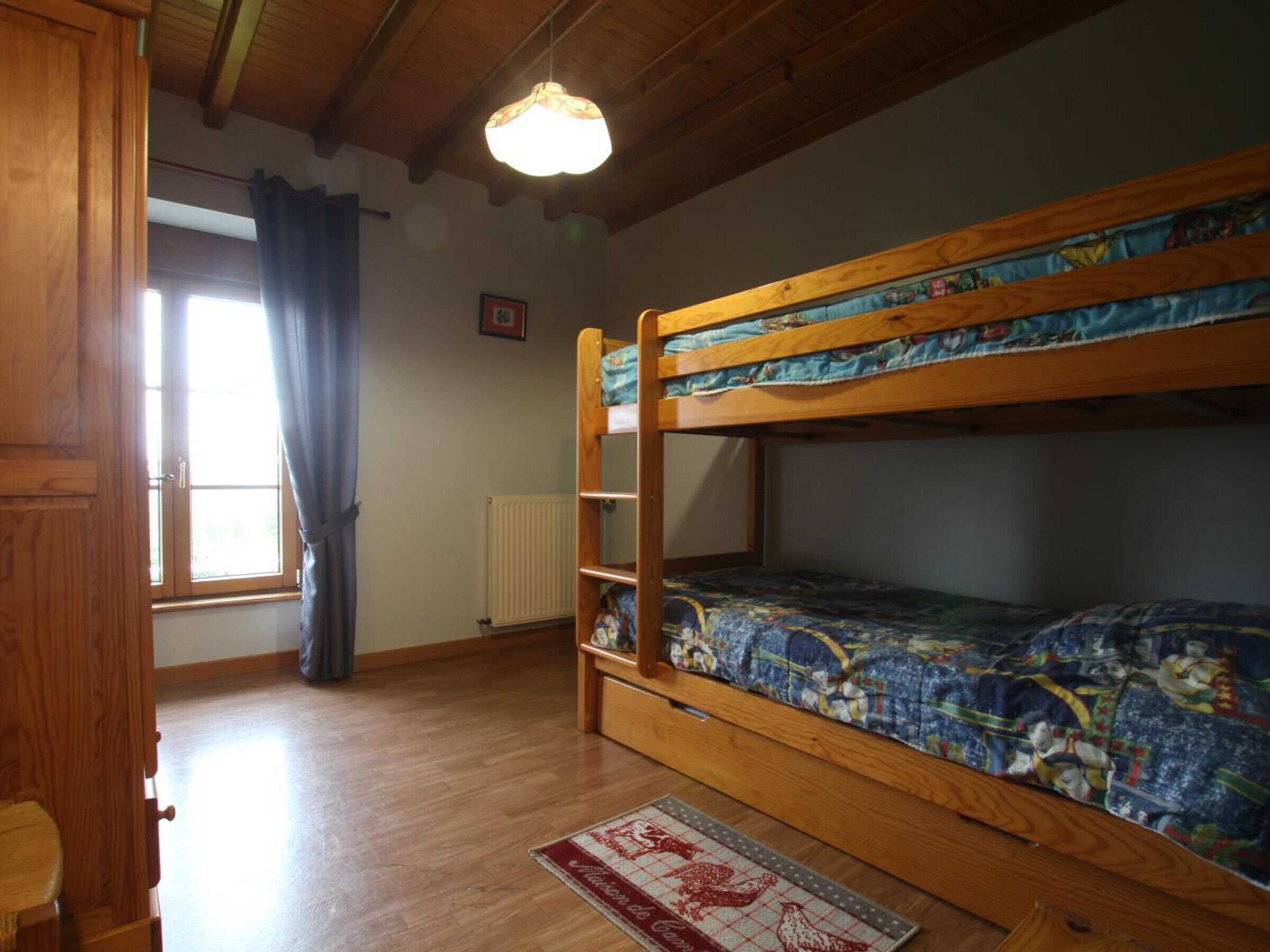 A room with a bunk bed, wooden flooring, a radiator, a window with curtains, and a door.