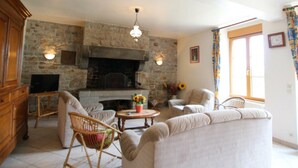 A living room with a stone fireplace, wicker furniture, and a large window with curtains. - Countryside house in Saussey, 4 bedrooms, peace and comfort (Beslon)