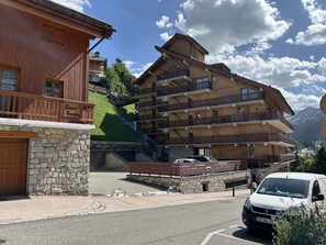 A multi-story wooden building with balconies, a stone facade, and a parking area with a white van.