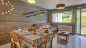 Un coin repas chaleureux, avec une table en bois dressée pour quatre personnes, entourée de chaises en bois. Un banc rembourré est placé contre le mur, et une petite table avec un livre se trouve à proximité. La pièce est dotée d’une grande fenêtre offrant une vue sur un paysage verdoyant.